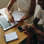 Duas pessoas sentadas em uma mesa de madeira analisam documentos financeiros ao lado de um notebook aberto. Uma das pessoas segura folhas impressas enquanto a outra utiliza a calculadora do smartphone. Sobre a mesa também há um bloco de anotações, canetas e outros itens de escritório. O ambiente interno é iluminado por luz natural suave, destacando uma cena de planejamento financeiro e organização de despesas.