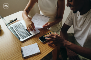 Duas pessoas sentadas em uma mesa de madeira analisam documentos financeiros ao lado de um notebook aberto. Uma das pessoas segura folhas impressas enquanto a outra utiliza a calculadora do smartphone. Sobre a mesa também há um bloco de anotações, canetas e outros itens de escritório. O ambiente interno é iluminado por luz natural suave, destacando uma cena de planejamento financeiro e organização de despesas.